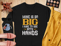 The Garment Graphics black shirt boldy states Mine Is So Big I Have To Use Both Hands in yellow and white. Its showcased on a wooden surface with makeup brushes, sunglasses, a yellow jacket, pink shoes, and makeup items.