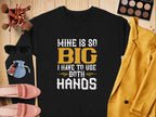 The Garment Graphics black shirt boldy states Mine Is So Big I Have To Use Both Hands in yellow and white. Its showcased on a wooden surface with makeup brushes, sunglasses, a yellow jacket, pink shoes, and makeup items.