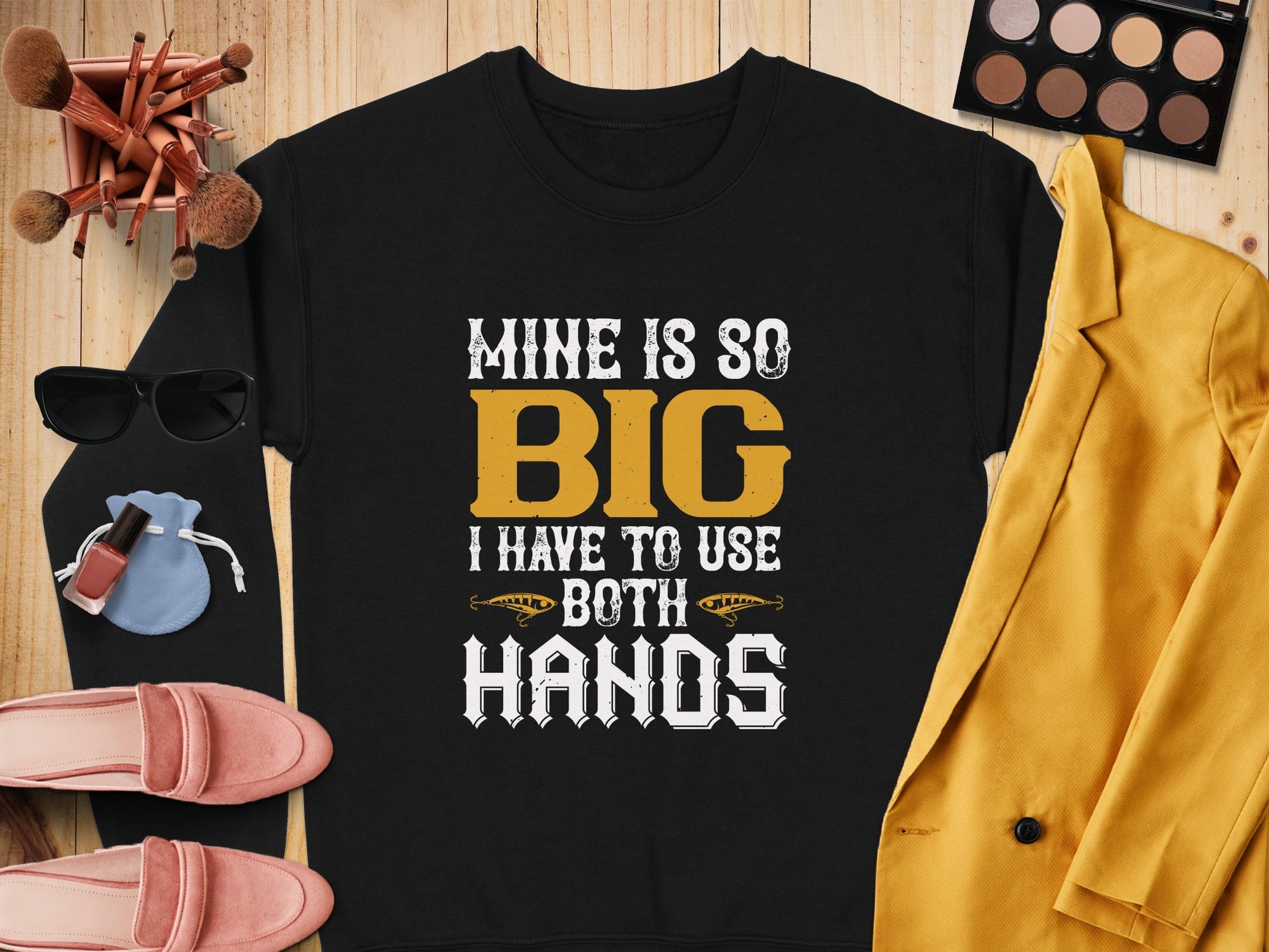 The Garment Graphics black shirt boldy states Mine Is So Big I Have To Use Both Hands in yellow and white. Its showcased on a wooden surface with makeup brushes, sunglasses, a yellow jacket, pink shoes, and makeup items.