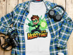 A Garment Graphics white sweatshirt with an Its Herotime superhero cartoon and vibrant green splashes is styled over a blue plaid shirt. Nearby, headphones, a camera, and a white coffee cup sit on the wooden surface—ideal for adventure-loving youth.