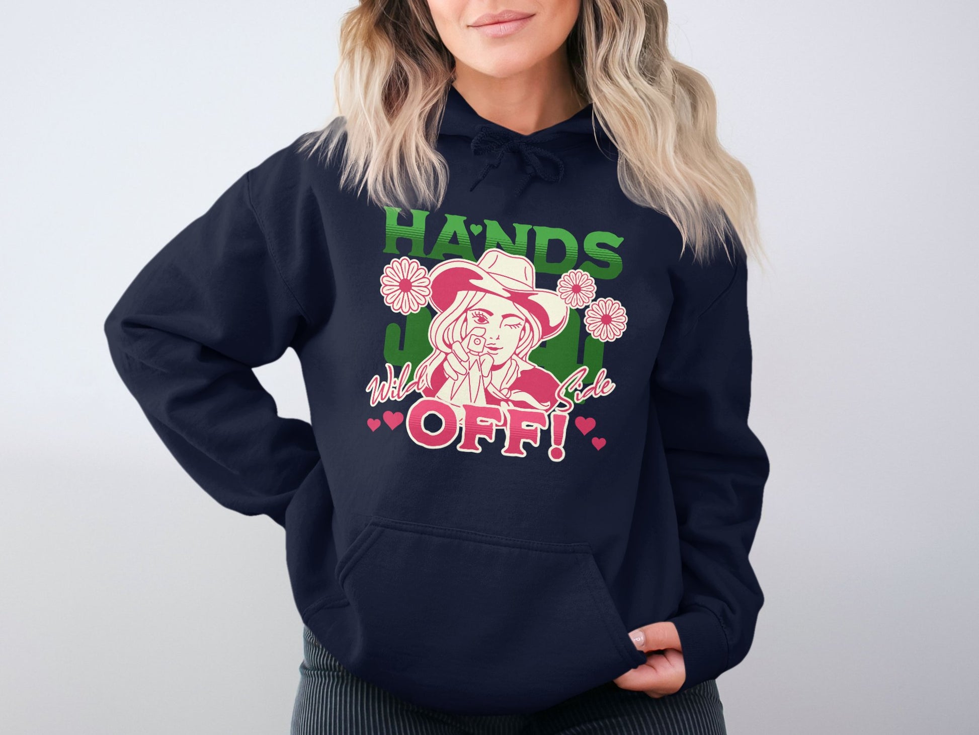 A person is seen wearing a navy Garment Graphics Bold Statement Hoodie featuring an illustration of a woman in a hat pointing, surrounded by flowers and hearts, with the text Hands Off, Wild Girl. Their wavy hair contrasts nicely with the plain wall backdrop.