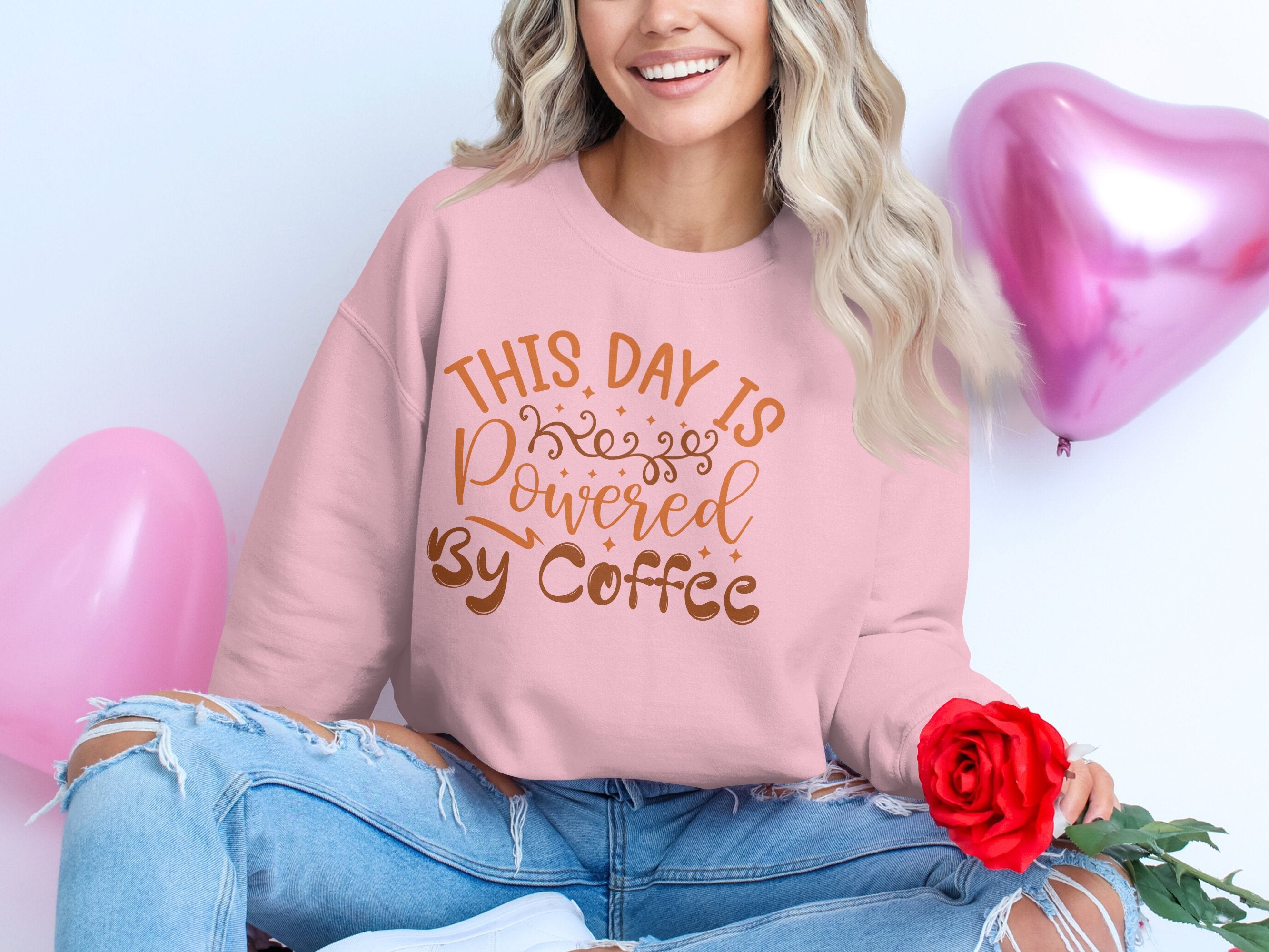 A woman wearing a Garment Graphics sweatshirt that says This Day is Powered by Coffee sits with a red rose, surrounded by pink heart balloons. Her long wavy hair and ripped jeans complete her casual style.
