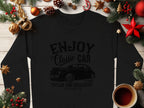 A black sweatshirt from Garment Graphics, featuring Enjoy Classic Car text and a vintage ride illustration, is displayed on a wooden table surrounded by pine branches, red and gold ornaments, dried orange slices, and a cozy coffee cup.