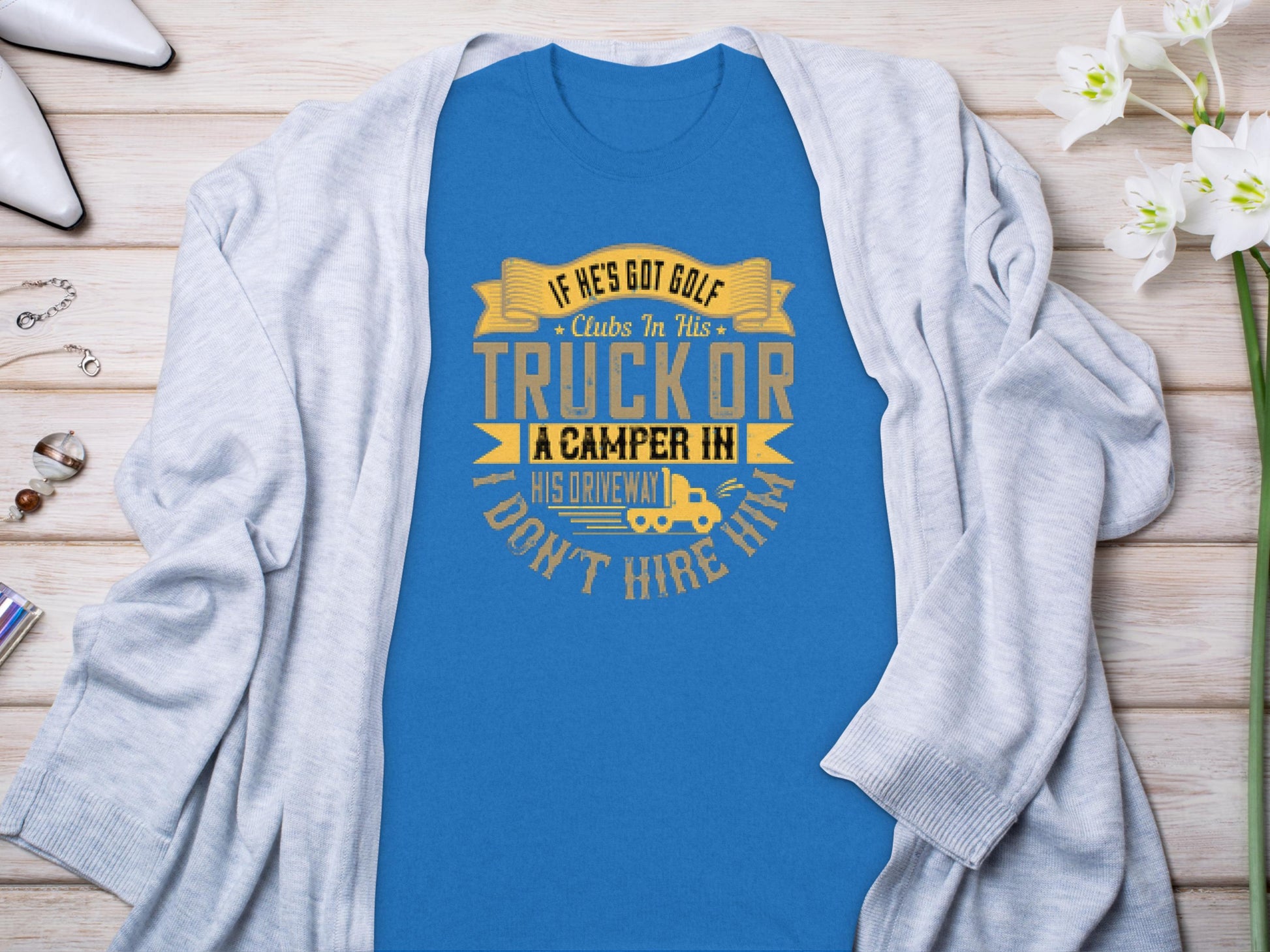 A Garment Graphics blue t-shirt with the funny graphic “If he’s got golf clubs in his truck or a camper in his driveway, don’t hire him,” styled on a gray cardigan surrounded by white flowers, jewelry, and shoes—ideal for those who appreciate camping humor.