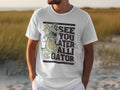 A person sports a Garment Graphics casual T-shirt with a cartoon alligator holding a basketball and the humorous text See You Later Alligator. The background showcases a beach scene with sand and grass, capturing leisurely vibes.