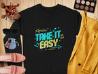 Garment Graphics presents a black unisex sweatshirt with Take It Easy in colorful text on a wooden surface, surrounded by a yellow jacket, pink shoes, sunglasses, makeup brushes and palette, nail polish, and a small blue pouch—ideal for casual wear.