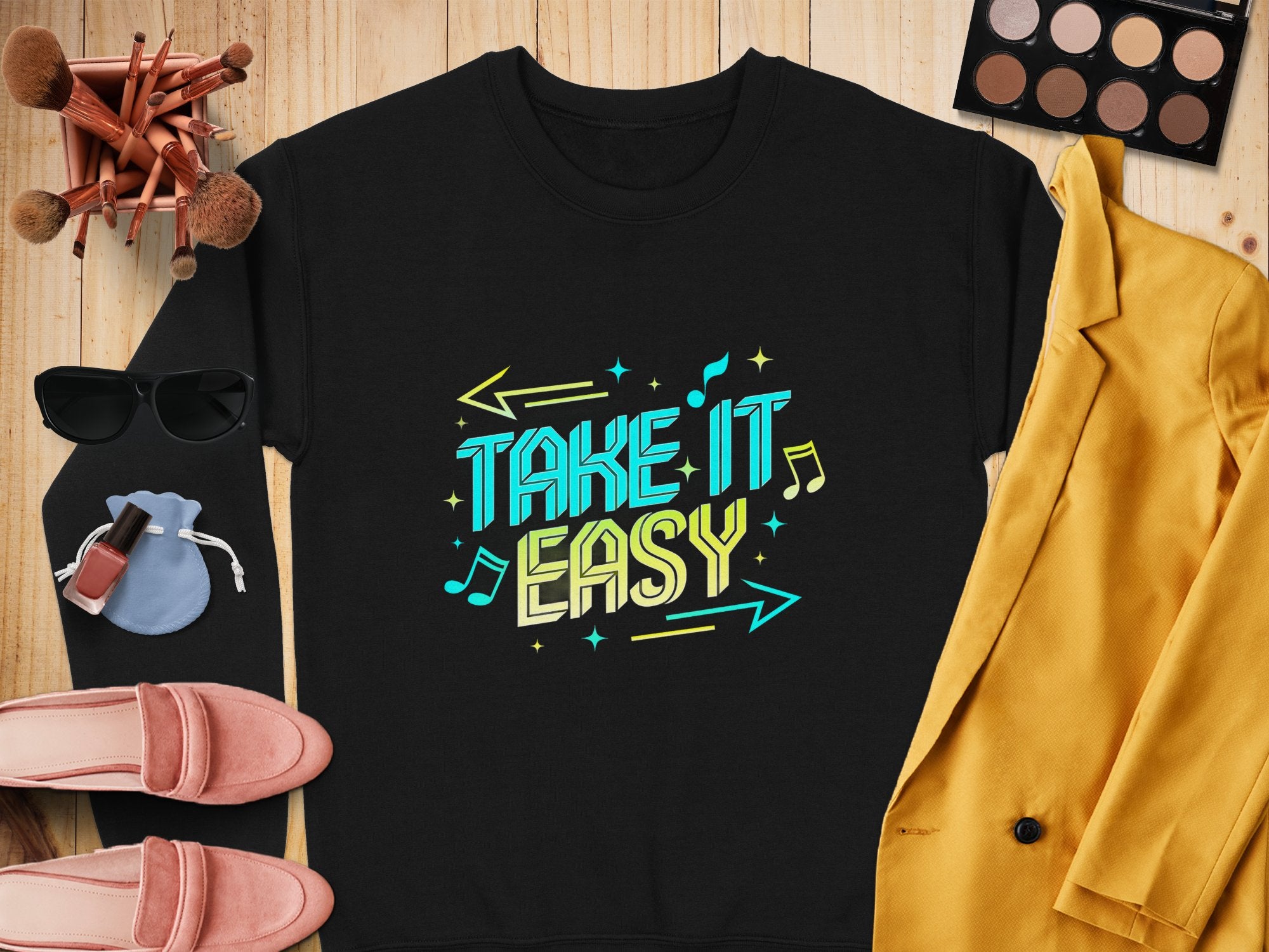 Garment Graphics presents a black unisex sweatshirt with Take It Easy in colorful text on a wooden surface, surrounded by a yellow jacket, pink shoes, sunglasses, makeup brushes and palette, nail polish, and a small blue pouch—ideal for casual wear.