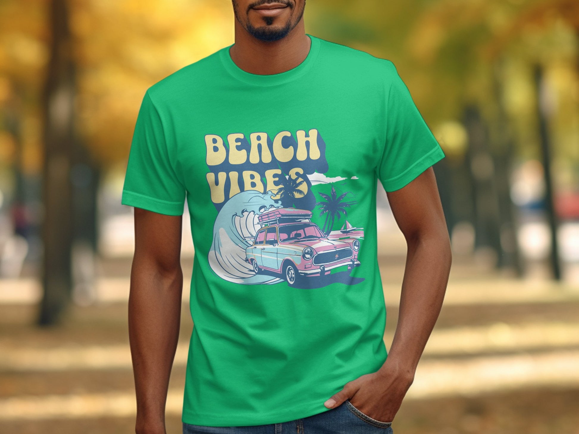 A person in a green Garment Graphics T-shirt with the text Beach Vibes displays classic cars, surfboards, palm trees, and a wave. The background features an autumnal park with blurred, warm-toned trees.