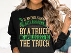 A person with long hair wears a Garment Graphics black unisex sweatshirt featuring a bold, humorous design with green and beige text: If an Englishman gets run down by a truck, he apologizes to the truck.