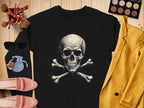 A Garment Graphics black shirt with a skull and crossbones design rests on wood, radiating edgy style. It is surrounded by makeup brushes, sunglasses, a pink purse, nail polish, pink shoes, an eyeshadow palette, and a vibrant yellow jacket.