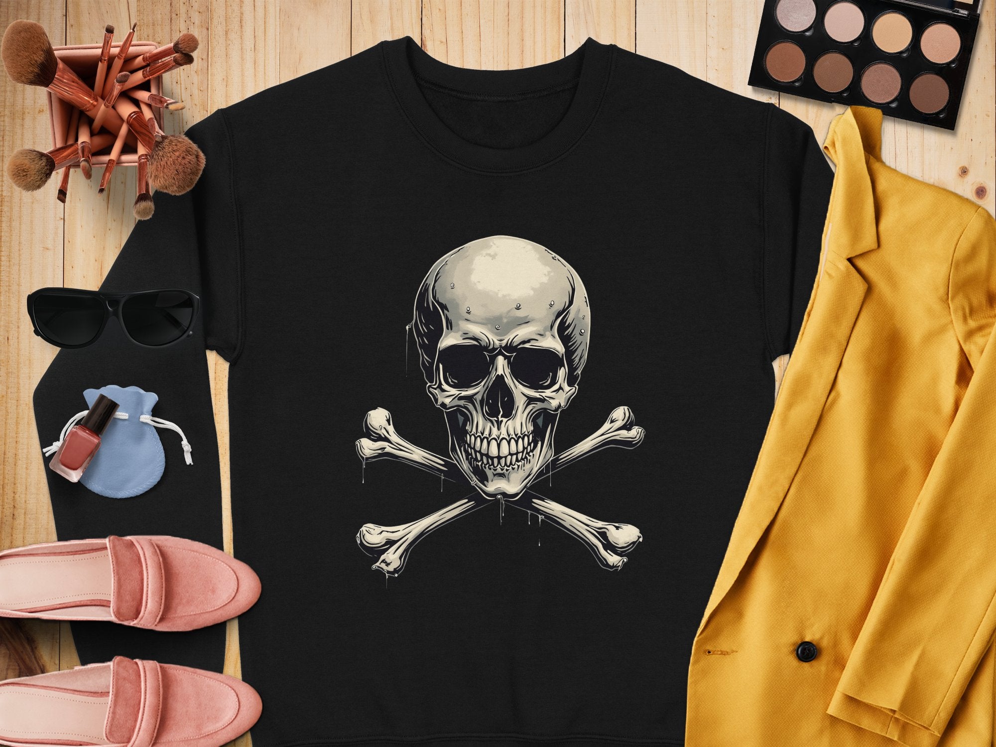 A Garment Graphics black shirt with a skull and crossbones design rests on wood, radiating edgy style. It is surrounded by makeup brushes, sunglasses, a pink purse, nail polish, pink shoes, an eyeshadow palette, and a vibrant yellow jacket.