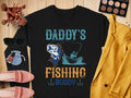 Black Garment Graphics shirt features a fishing illustration with Daddys Fishing Buddy text, ideal for fishing enthusiasts. Surrounded by makeup brushes, sunglasses, pink shoes, nail polish, eyeshadow palette, and a yellow jacket on a wooden background.