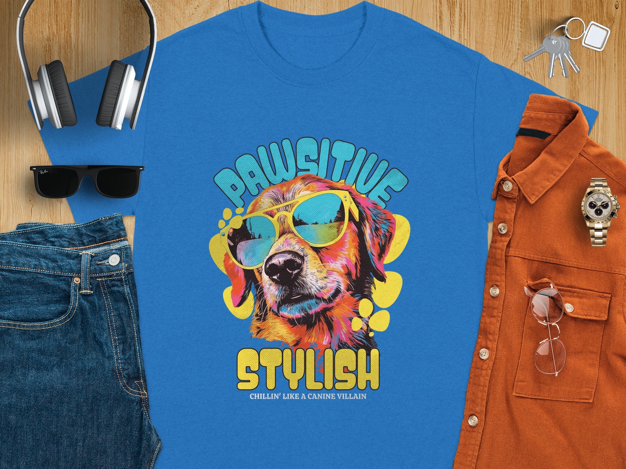 A Garment Graphics blue dog t-shirt features a pooch in sunglasses with Paw-sitive Stylish, Chillin Like A Canine Villain, styled with jeans, shades, headphones, a watch, and a brown shirt on a wooden surface.