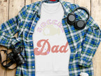 A flat lay image features a blue and green plaid shirt next to Garment Graphics Soccer Dad sweatshirt with a soccer ball design. A camera, headphones, and white travel mug enhance the casual vibe on the wooden surface—ideal for fans of unisex sizing.