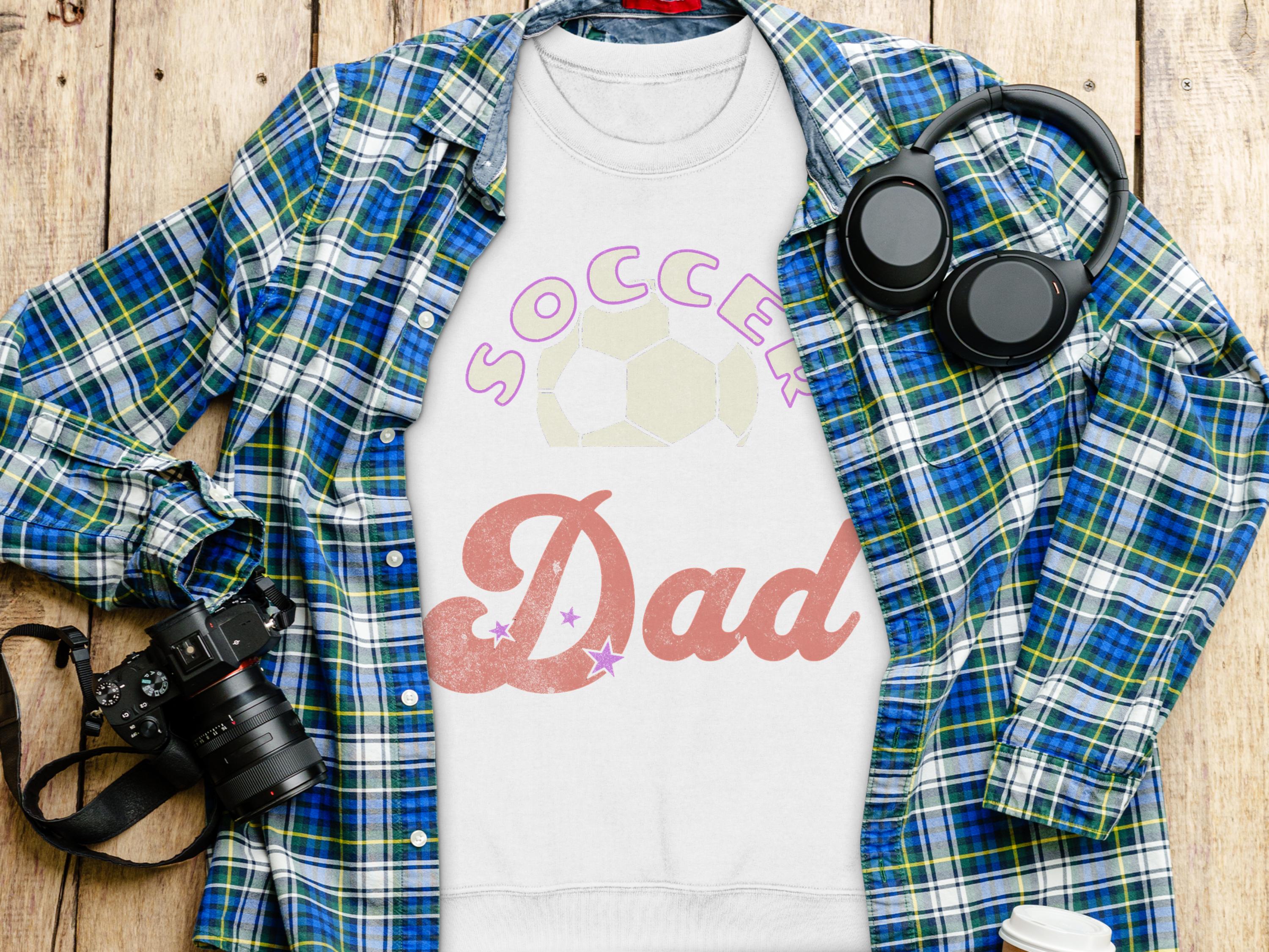 A flat lay image features a blue and green plaid shirt next to Garment Graphics Soccer Dad sweatshirt with a soccer ball design. A camera, headphones, and white travel mug enhance the casual vibe on the wooden surface—ideal for fans of unisex sizing.