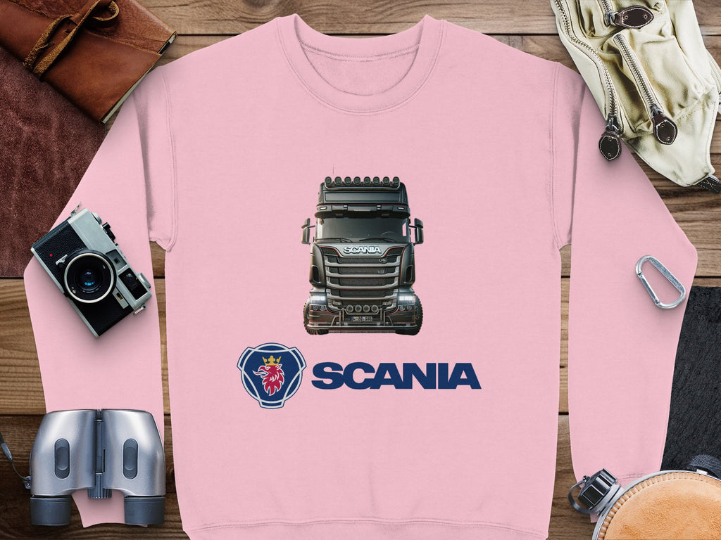 A pink Garment Graphics crewneck sweatshirt with a Scania truck and logo lies on a wooden surface, surrounded by a camera, binoculars, leather bag, goggles, and carabiner—ideal for truck enthusiasts.