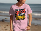 A person wears a vibrant Garment Graphics Its a Good Day to Laugh pink t-shirt featuring red, white, and yellow letters while standing on the beach with a softly blurred ocean background.