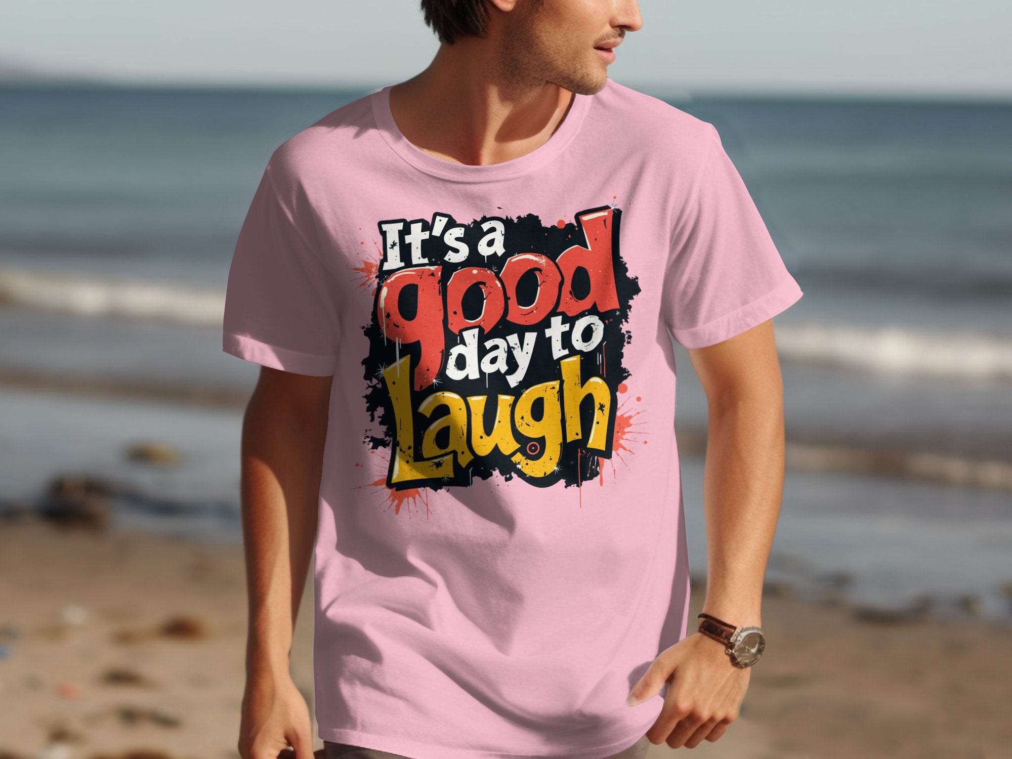 A person wears a vibrant Garment Graphics Its a Good Day to Laugh pink t-shirt featuring red, white, and yellow letters while standing on the beach with a softly blurred ocean background.