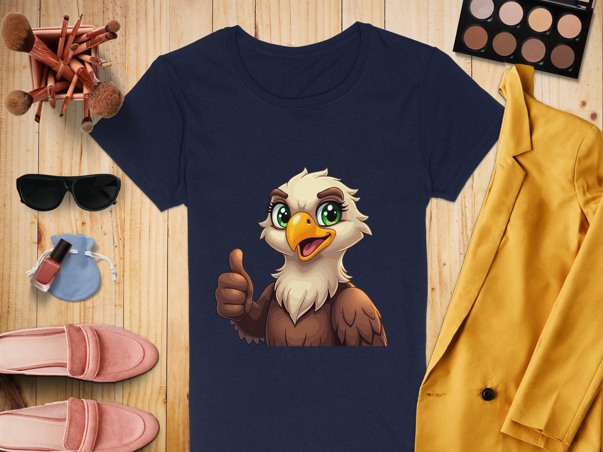 A Garment Graphics navy blue T-shirt with a cartoon eagle giving a thumbs-up is displayed on wood, surrounded by makeup brushes, a sunglasses case, yellow jacket, pink shoes, nail polish, and an eyeshadow palette—ideal for stylish animal lovers!.