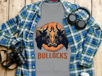 A gray T-shirt by Garment Graphics features a striking bullock design with two bulls facing each other and BULLOCKS in vivid orange. Displayed on a wooden surface, it complements a blue plaid shirt, headphones, camera, and white cup beautifully.