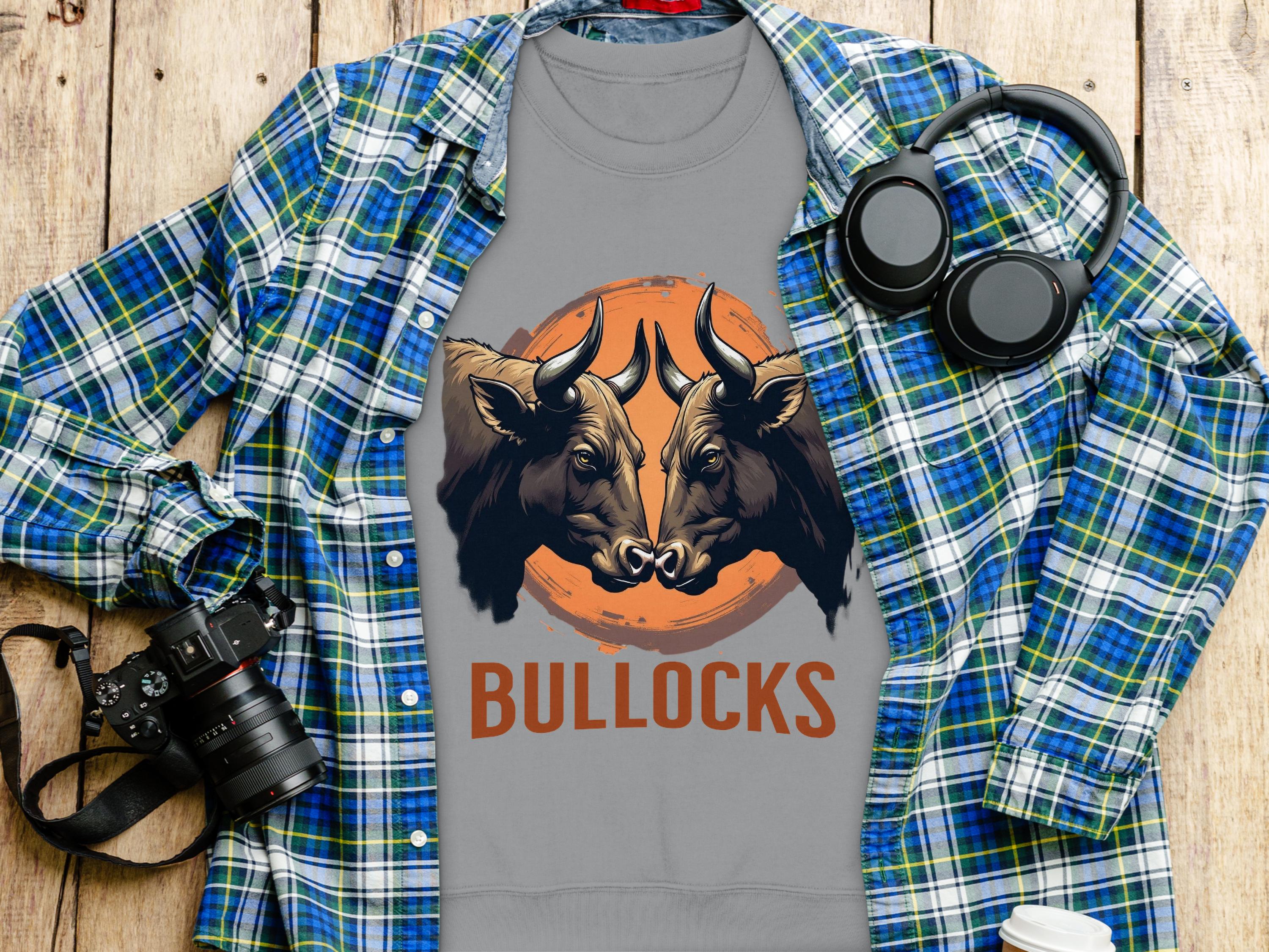 A gray T-shirt by Garment Graphics features a striking bullock design with two bulls facing each other and BULLOCKS in vivid orange. Displayed on a wooden surface, it complements a blue plaid shirt, headphones, camera, and white cup beautifully.