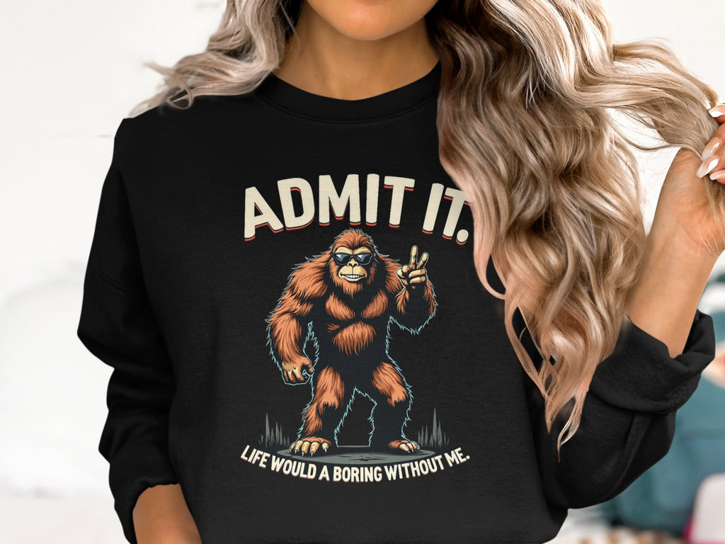 A person with long curly hair is wearing a black Garment Graphics sweatshirt featuring Bigfoot and the text, ADMIT IT. Life would be boring without me.