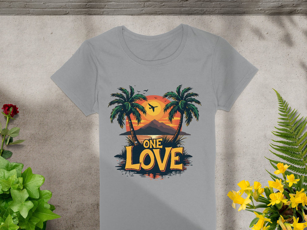 Garment Graphics gray T-shirt displays One Love under a Tropical Sunset with palm trees and a plane. Laid flat on concrete amidst vibrant green and yellow plants, its perfect for your next beach outing.