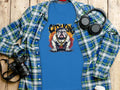 A blue t-shirt from Garment Graphics with a bulldog and gold chains is placed under a blue plaid shirt, alongside black headphones, a camera, and a white paper cup on a wooden surface.