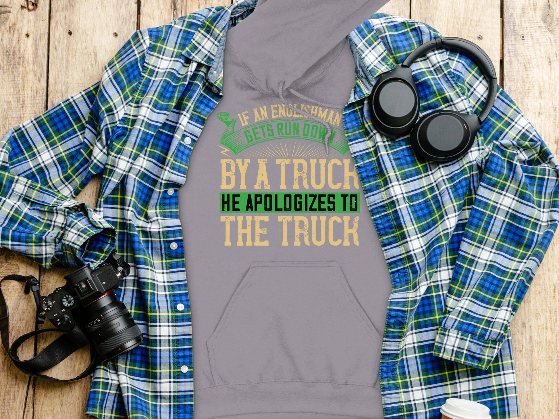 A plaid shirt, camera, headphones, and coffee cup surround a gray Garment Graphics hoodie featuring a funny quote about an engineer’s take on an accident, humorously apologizing to a truck.