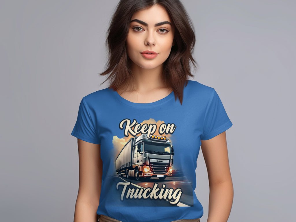 A brown-haired woman wears a Garment Graphics blue graphic T-shirt featuring a powerful truck and the bold text Keep On Trucking against a neutral gray background, highlighting the striking design.