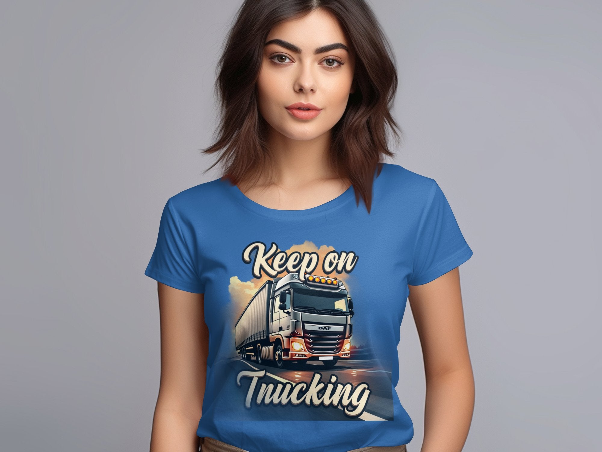 A brown-haired woman wears a Garment Graphics blue graphic T-shirt featuring a powerful truck and the bold text Keep On Trucking against a neutral gray background, highlighting the striking design.