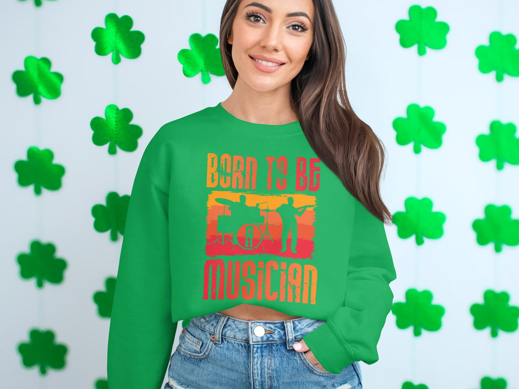 A woman joyfully poses before a wall of green shamrocks, wearing Garment Graphics unisex green sweater with Born to Be Musician emblazoned on it, celebrating her love for music. Her long hair frames her smiling face, showcasing her musical passion.