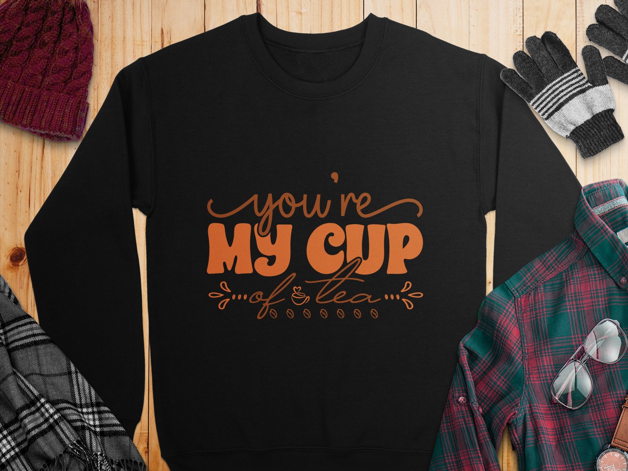 Garment Graphics presents a unisex sweatshirt with Youre My Cup of Tea in bold orange, creatively laid out with a plaid shirt, scarf, knit gloves, and maroon knit hat on a wooden surface.