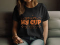 A person sits on a couch wearing a Garment Graphics black graphic T-shirt made of 100% cotton, featuring the bold orange text Youre My Cup of Tea, captured partially from the shoulders to the waist.