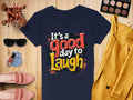 A navy blue, semi-fitted T-shirt from Garment Graphics, featuring a colorful Its a good day to laugh graphic print, is displayed on a wooden surface alongside sunglasses, makeup brushes, nail polish, a round purse, pink shoes, an eyeshadow palette, and a yellow blazer.