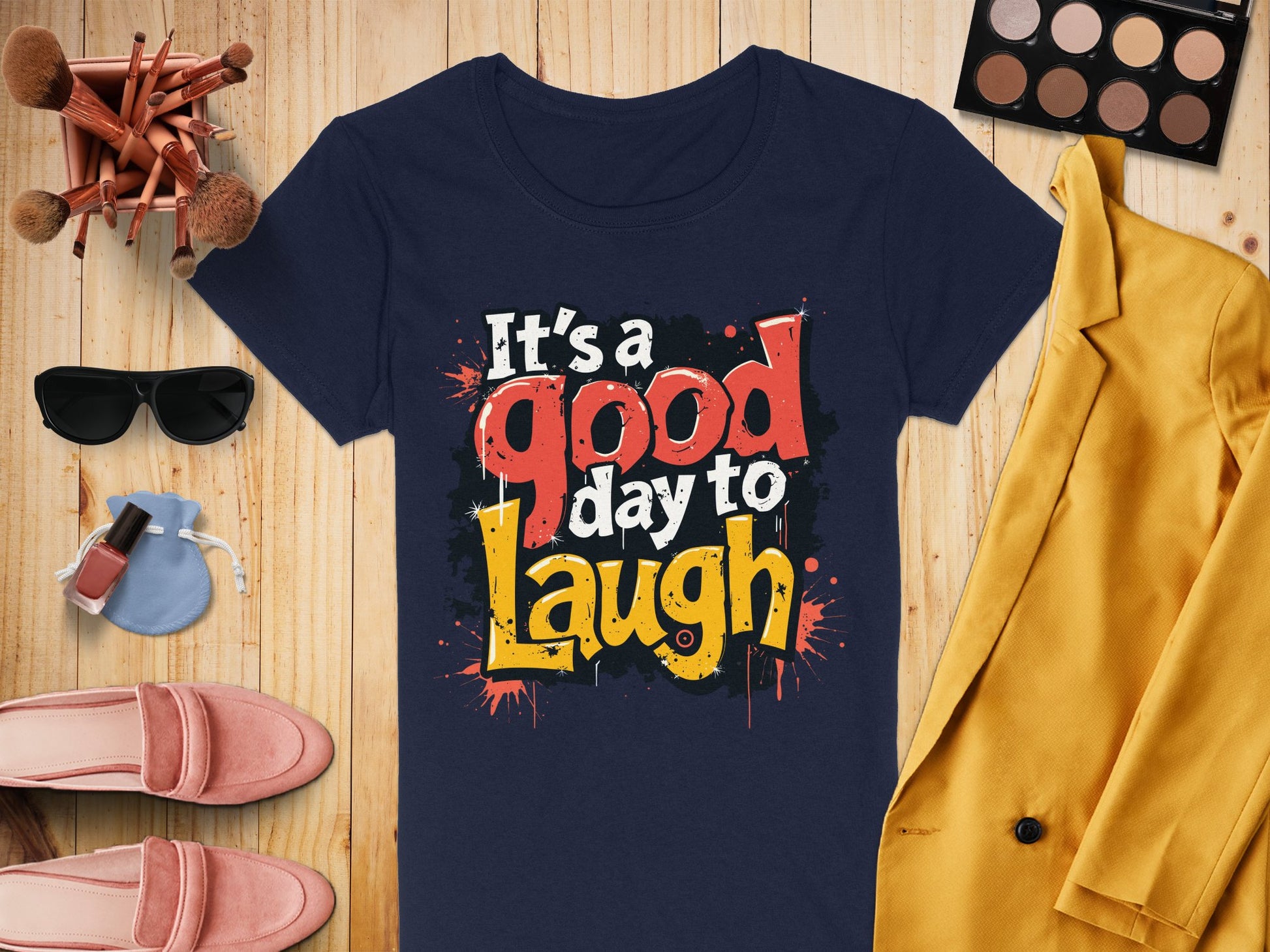 A navy blue, semi-fitted T-shirt from Garment Graphics, featuring a colorful Its a good day to laugh graphic print, is displayed on a wooden surface alongside sunglasses, makeup brushes, nail polish, a round purse, pink shoes, an eyeshadow palette, and a yellow blazer.