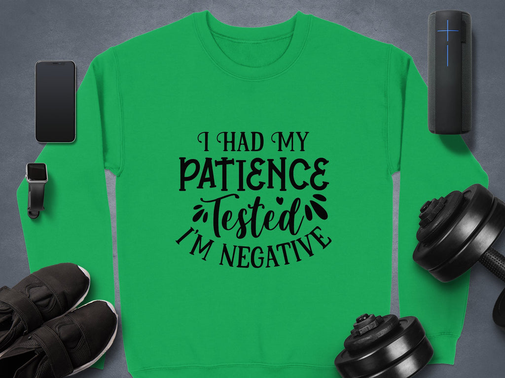 A green unisex sweatshirt by Garment Graphics, featuring I had my patience tested, Im negative, is laid flat. Its ideal for casual outings and rests on a gray textured background surrounded by a smartphone, smartwatch, headphones, portable speaker, and dumbbells.