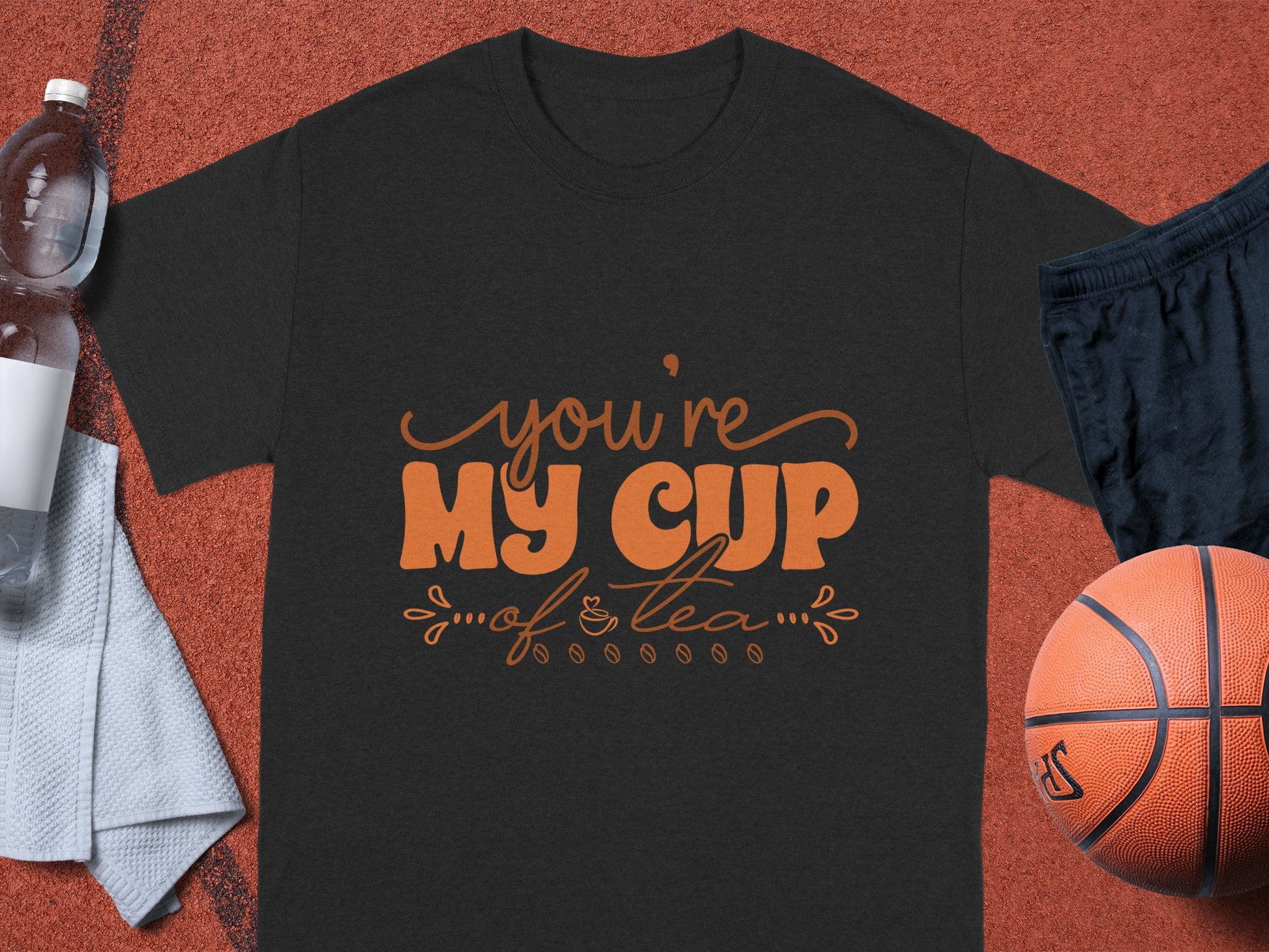 Garment Graphics 100% cotton black graphic T-shirt displays Youre My Cup of Tea in orange. Its placed on a bright red sports court alongside a basketball, black shorts, a towel, and a water bottle.