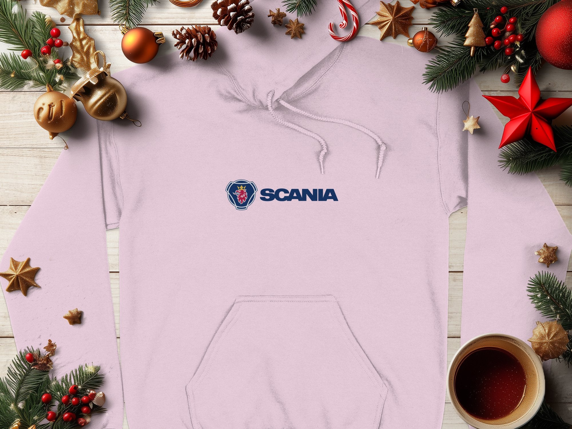 A Garment Graphics white hoodie with the blue Scania logo is displayed on a wooden surface. Its surrounded by festive pine branches, ornaments, pine cones, and a steaming cup of coffee, creating a cozy holiday atmosphere.