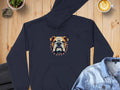 A Garment Graphics black hoodie showcasing a bold bulldog design with a spiked collar is on a wooden surface. Nearby items include a coffee cup featuring latte art, a denim shirt, dark pants, and a small plant in a pot.