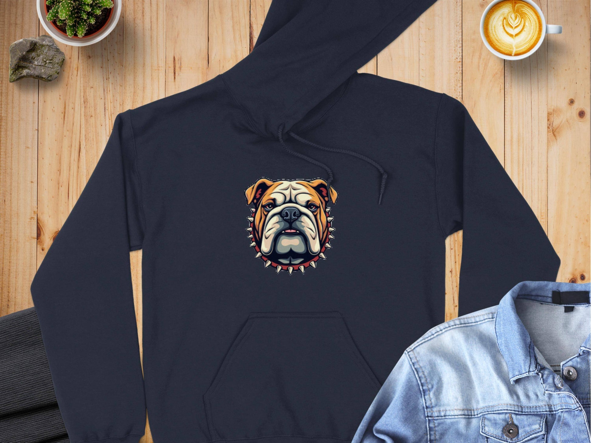 A Garment Graphics black hoodie showcasing a bold bulldog design with a spiked collar is on a wooden surface. Nearby items include a coffee cup featuring latte art, a denim shirt, dark pants, and a small plant in a pot.