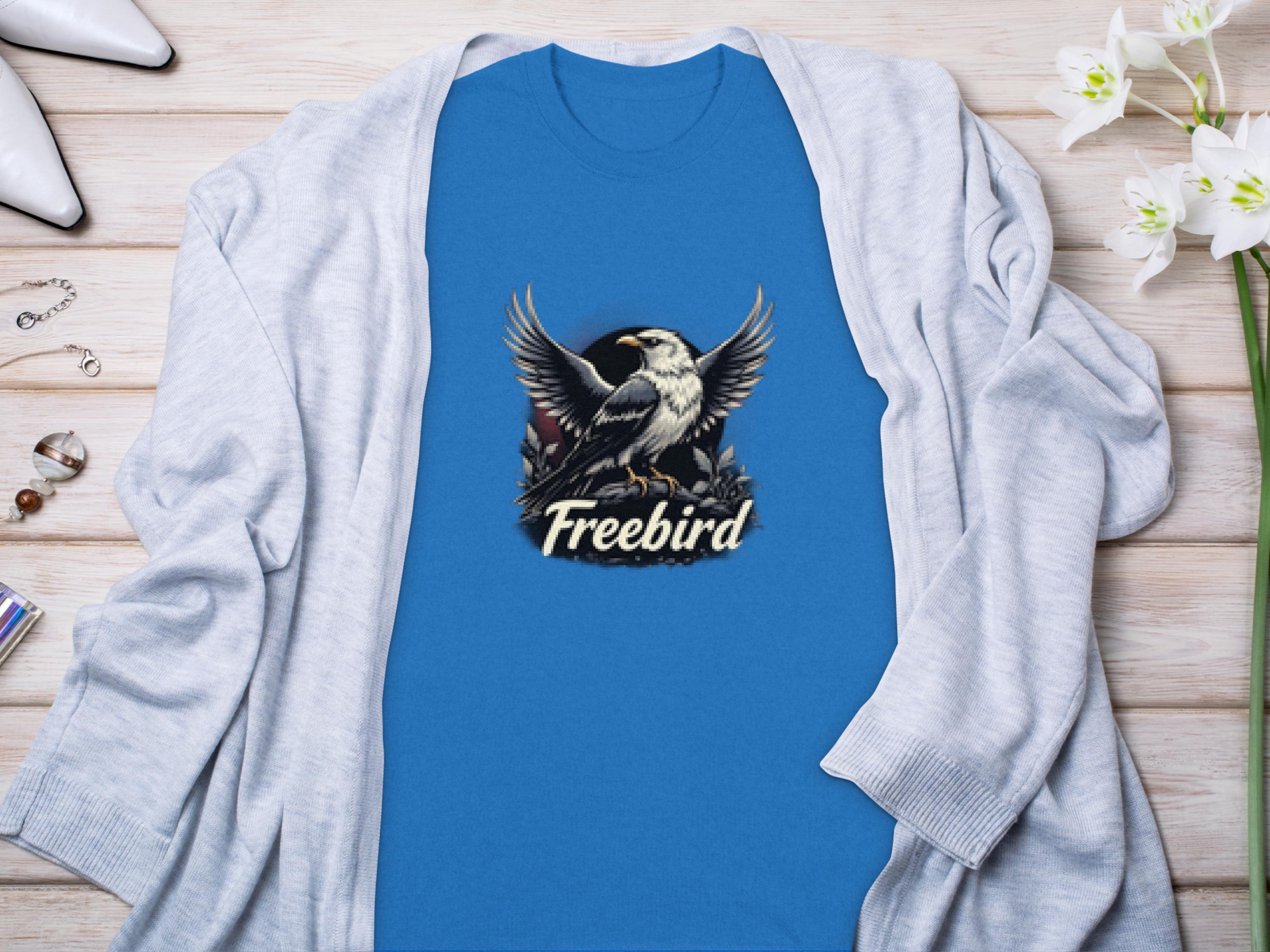 A blue T-shirt by Garment Graphics showcases a Freebird logo with an illustrated bird with outstretched wings. Ideal for your casual style, its displayed on wood alongside a light gray cardigan, jewelry, white shoes, and white flowers.
