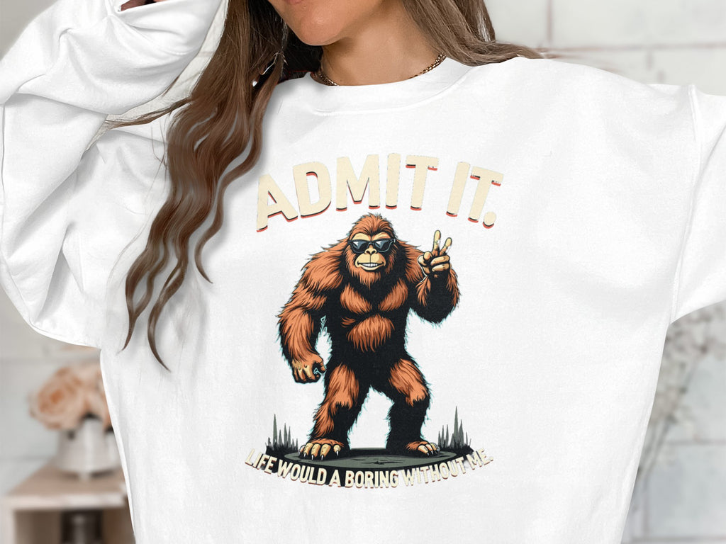 Someone in casual wear sports a Garment Graphics white sweatshirt featuring Bigfoot. The text reads, ADMIT IT above and LIFE WOULD B A BORING WITHOUT ME below. Their long brown hair enhances the relaxed fit provided by the unisex sizing.