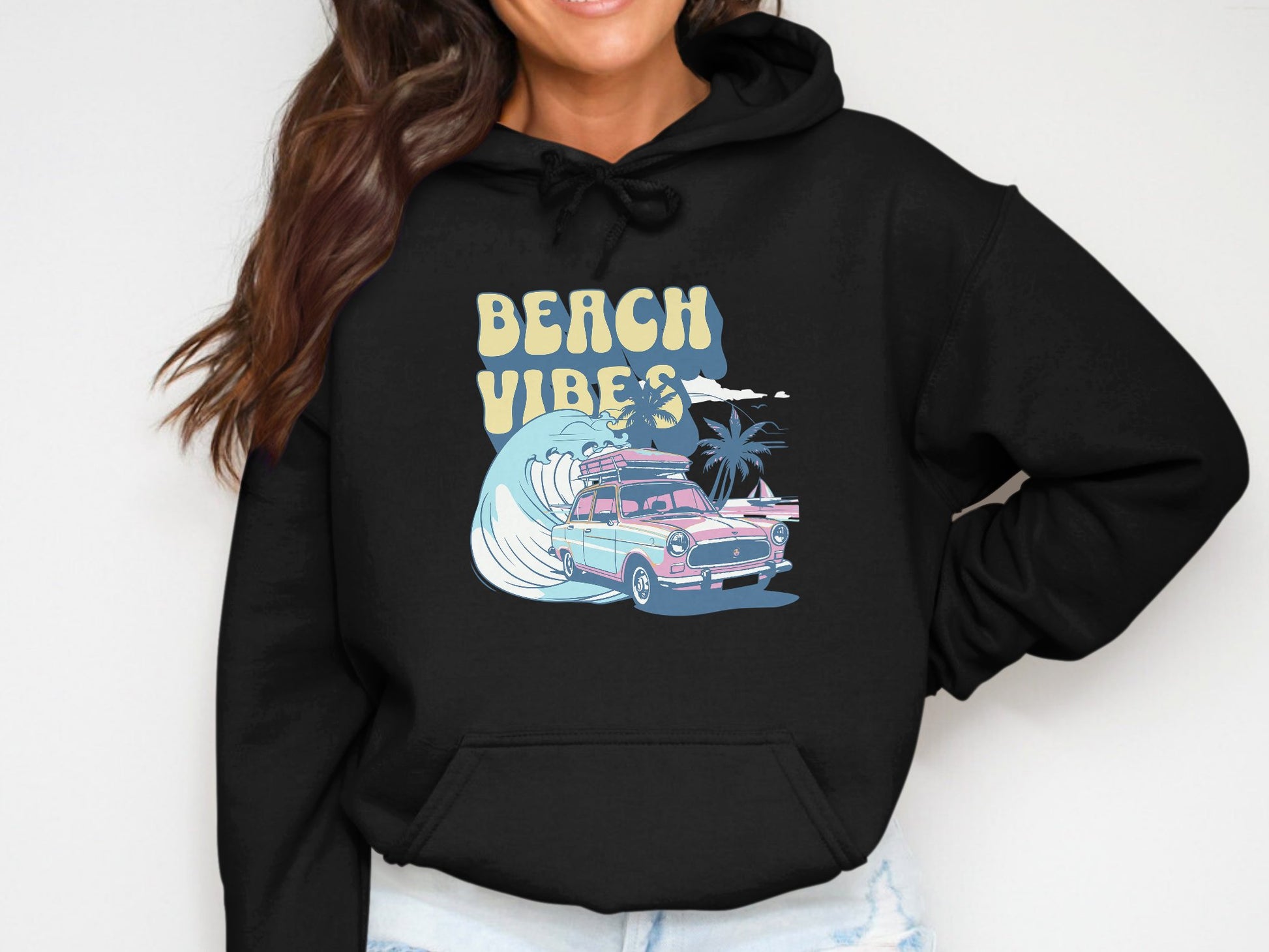 A person dons a vibrant black Garment Graphics hoodie featuring BEACH VIBES above a classic car and surfboard. The background boasts retro beach flair with a large wave, palm trees, and a seagull.