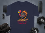 A navy blue Garment Graphics T-shirt features a cartoon turkey running above TURKEY ON THE RUN. The gray surface around the shirt displays a smartphone, smartwatch, wireless speaker, dumbbell, and black sneakers.