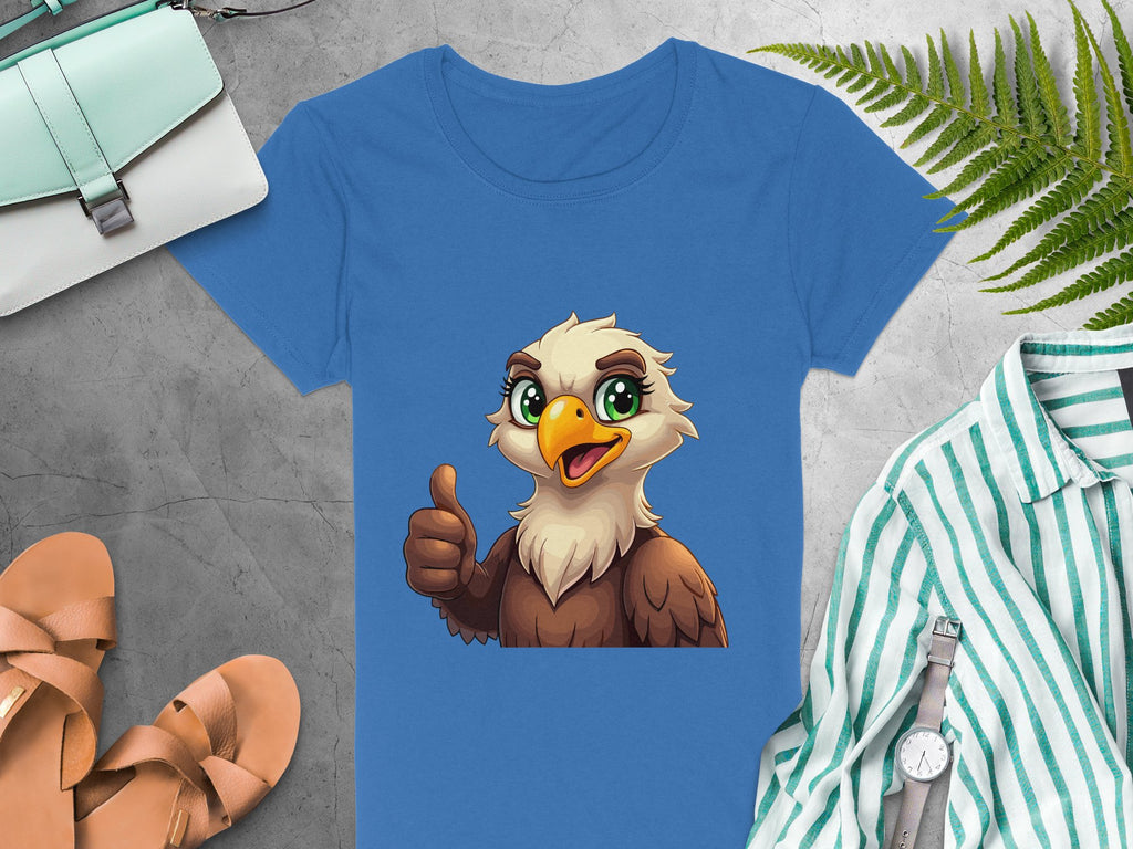 The Garment Graphics Blue Eagle T-shirt showcases a cartoon eagle giving a thumbs-up. Its paired with a green bag, fern leaves, tan sandals, striped shirt, and watch on a gray stone surface. Ideal for animal lovers.