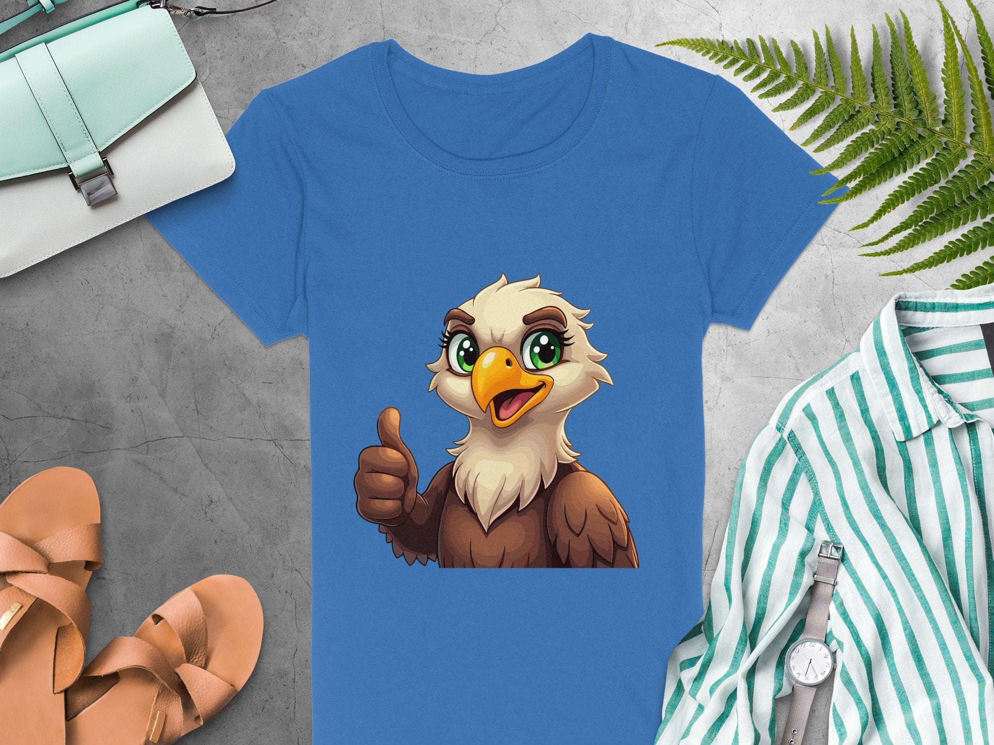 The Garment Graphics Blue Eagle T-shirt showcases a cartoon eagle giving a thumbs-up. Its paired with a green bag, fern leaves, tan sandals, striped shirt, and watch on a gray stone surface. Ideal for animal lovers.