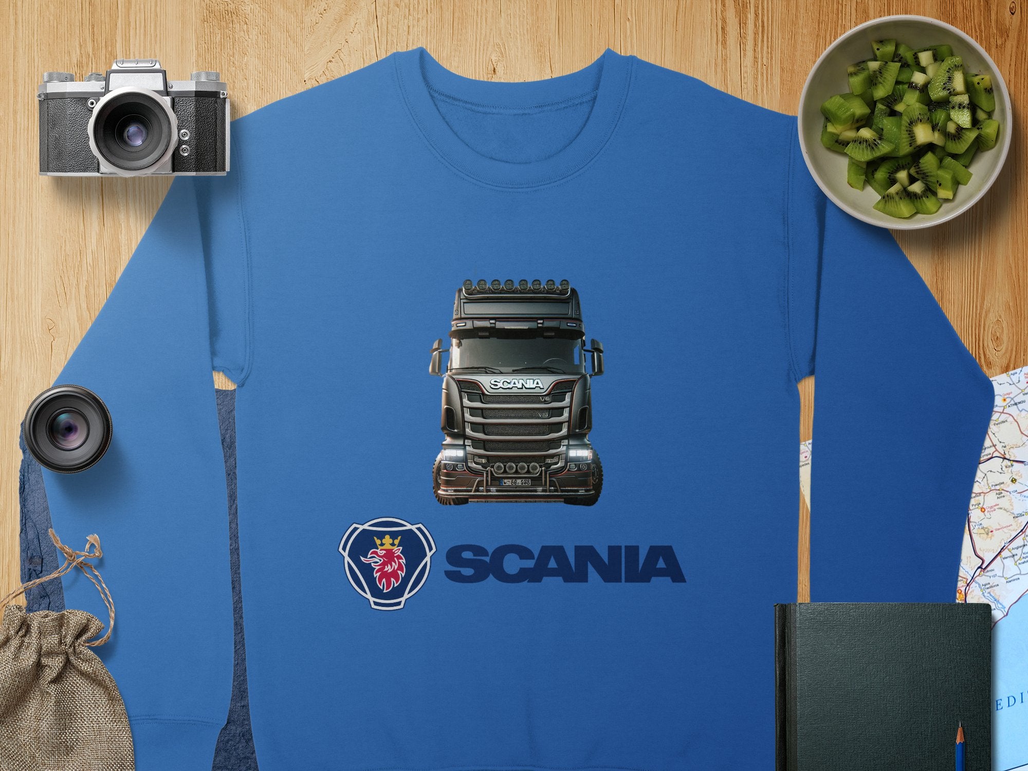 Garment Graphics offers a blue crewneck sweatshirt displaying a Scania truck prominently above the logo, ideal for truck enthusiasts. The product is stylishly surrounded by a camera, lens, fruit bowl, notebook, pouch, and map on a wooden surface.