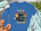 A Garment Graphics blue graphic sweatshirt with a green truck and Keep on Trucking text rests on a gray surface. Ideal for truck lovers, its paired with a green bag, brown sandals, striped shirt, watch, and fern leaves.