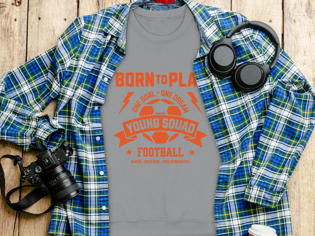 A gray Garment Graphics T-shirt featuring BORN TO PLAY and YOUNG SQUAD FOOTBALL in orange, laid on wood, pairs with a blue plaid shirt, black headphones, and a camera. Perfect for young squads, its unisex sizing suits any football enthusiasts wardrobe.